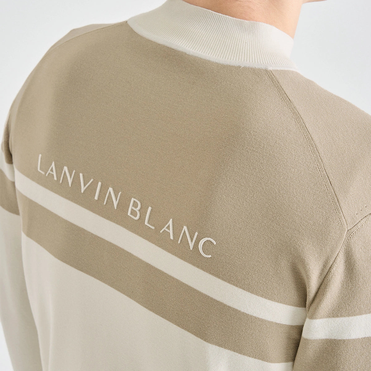 Close-up of the back of a beige knit sweater with a white ribbed mock neck, horizontal white stripe, and 'LANVIN BLANC' text printed across the upper back.