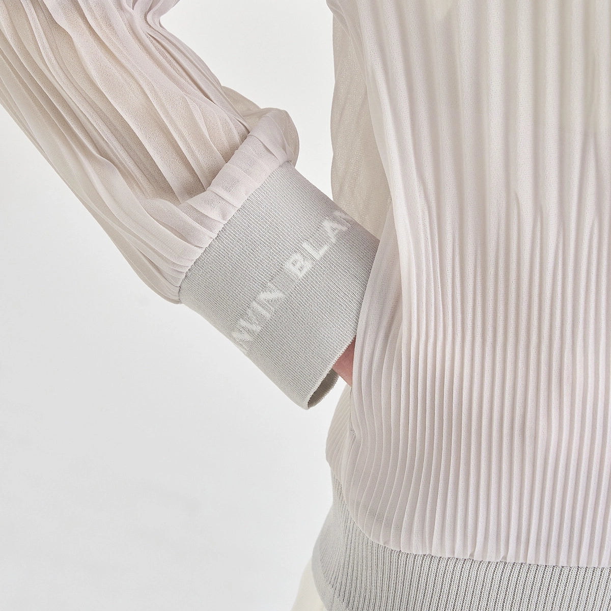 Close-up of a white pleated long-sleeve top with a ribbed cuff, showing the sleeve cuff and part of the torso against a light studio background.