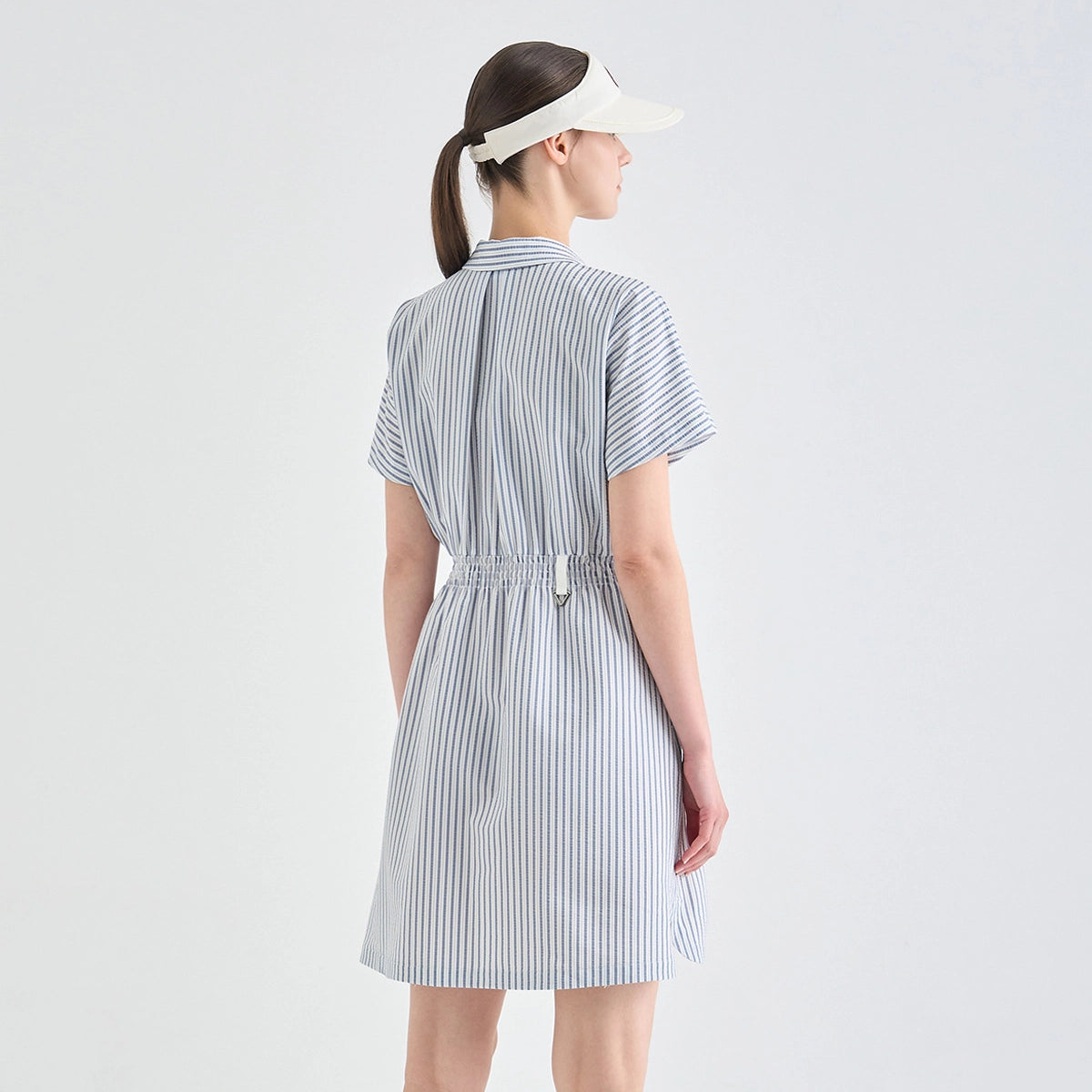 A woman shown from the back wearing a short-sleeve striped dress with a tie waist and a white visor, photographed against a plain light gray studio background.