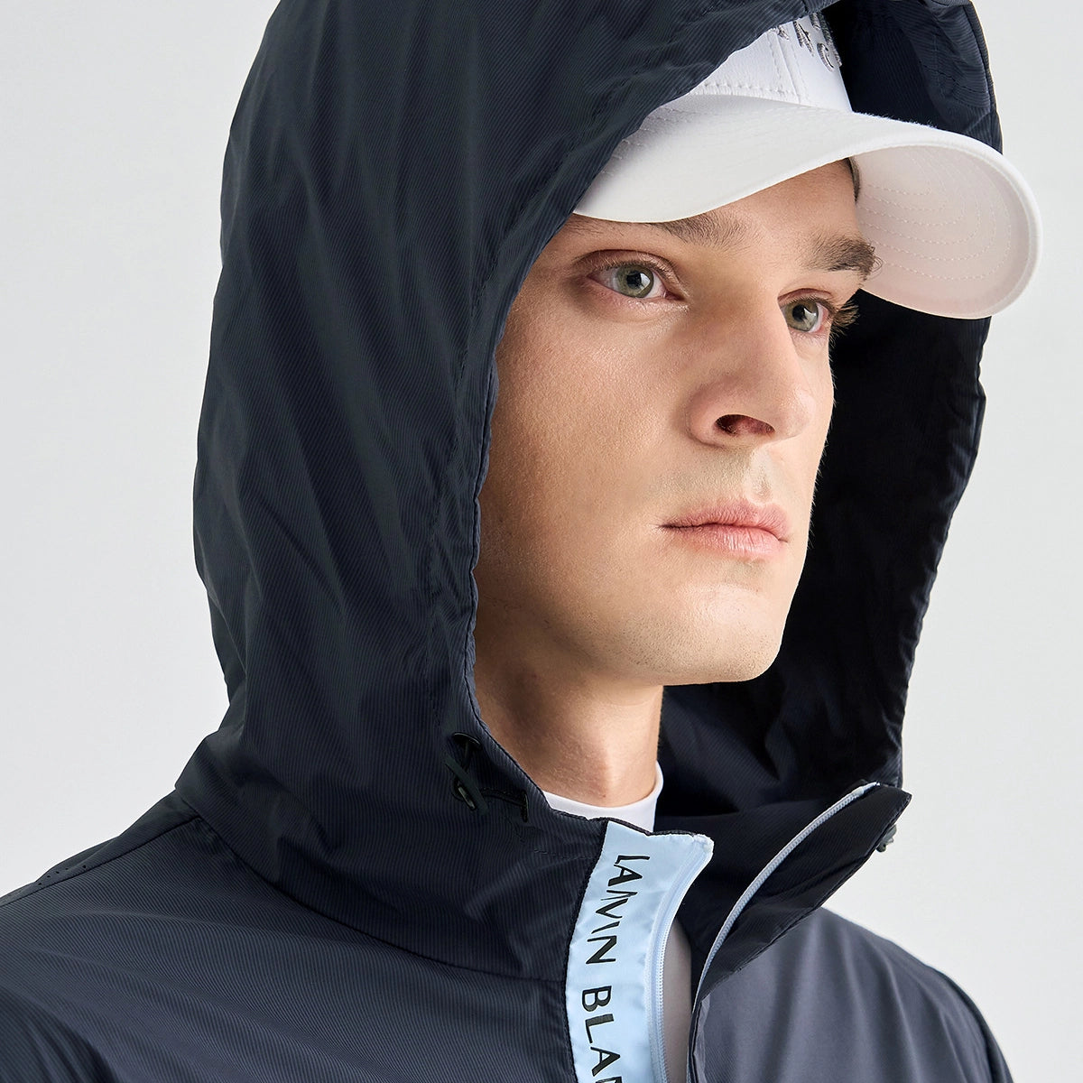 A close-up of a man wearing a dark navy hooded jacket with a white visor cap and a white t-shirt underneath. The jacket has a light blue inner lining near the collar with black text reading 'LANVIN BLANC'.