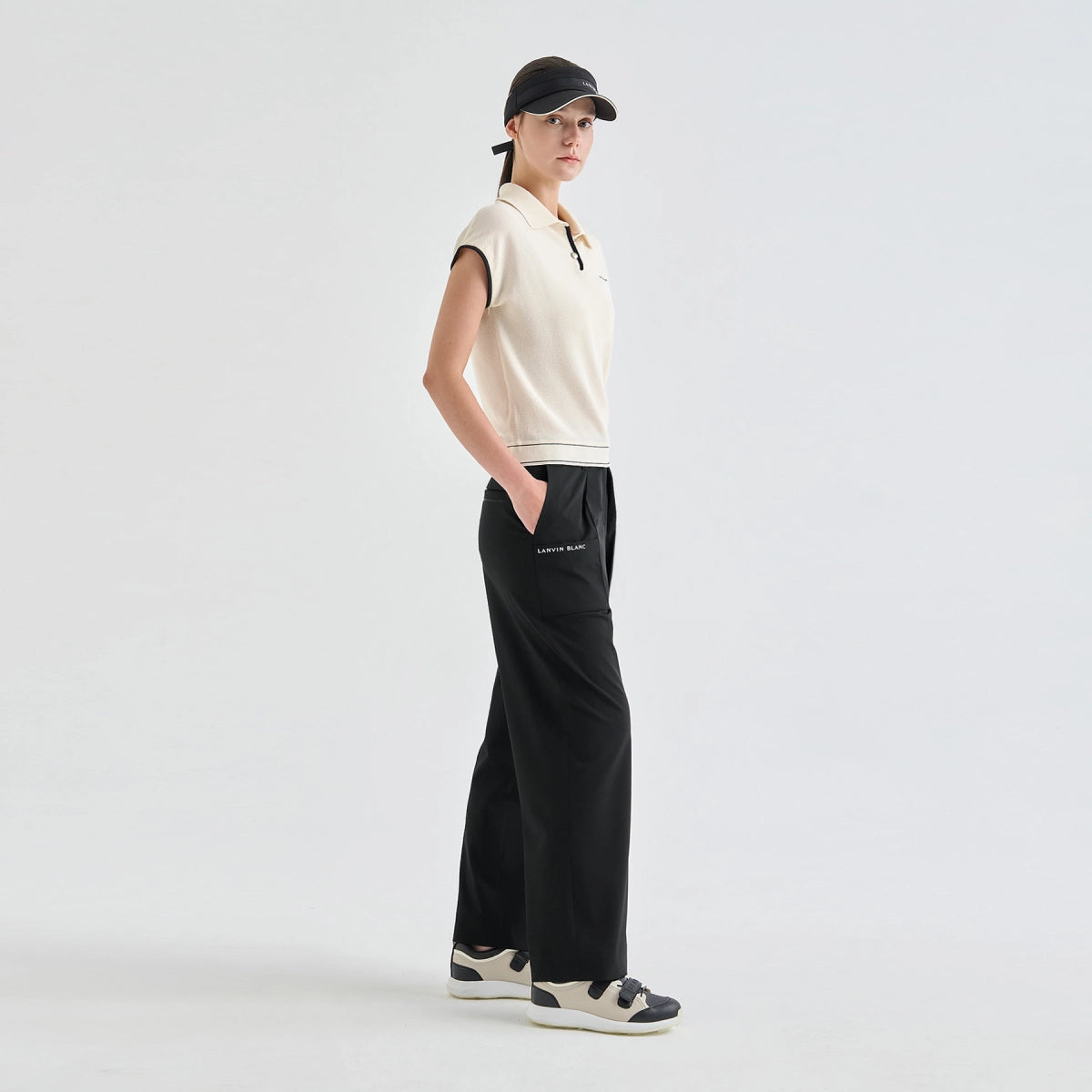 A model stands in profile wearing a cream sleeveless knit polo shirt, black wide-leg pants with a small white logo on the pocket, and black-and-white sneakers against a clean light gray studio background.