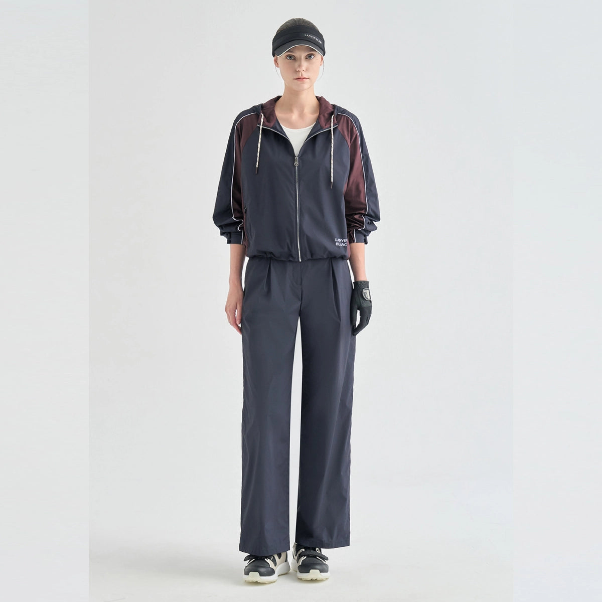 A model stands against a light gray studio background wearing a dark navy athletic tracksuit with burgundy hooded jacket panels, a white undershirt, black gloves, and a black cap.