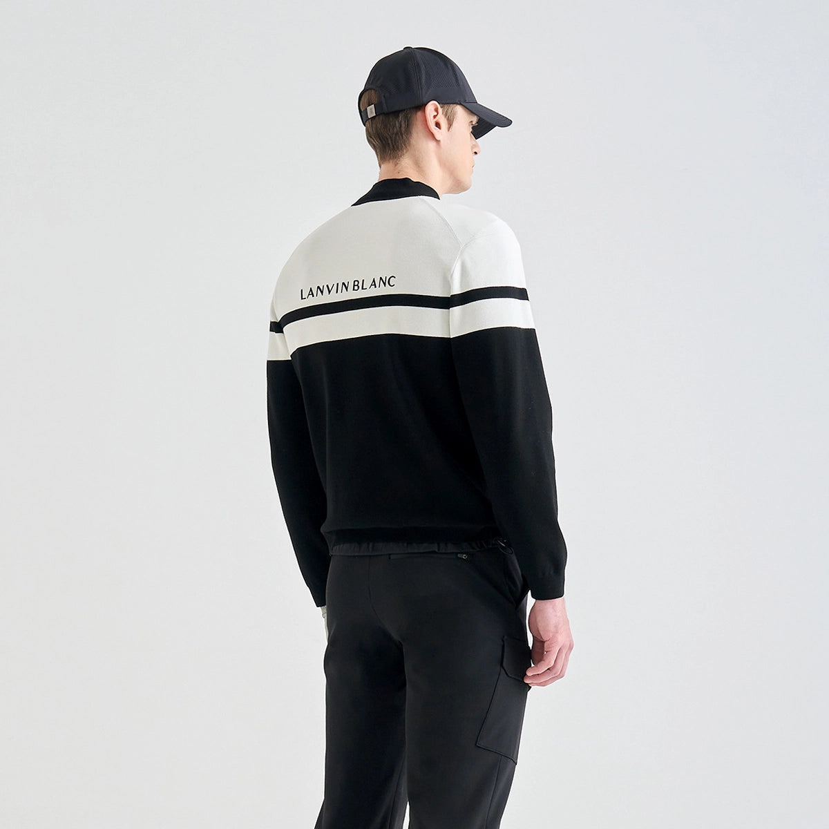 A man wearing a black and white color-blocked Lanvin Blanc track jacket, black trousers, and a black baseball cap, shown from the back against a light gray studio background.