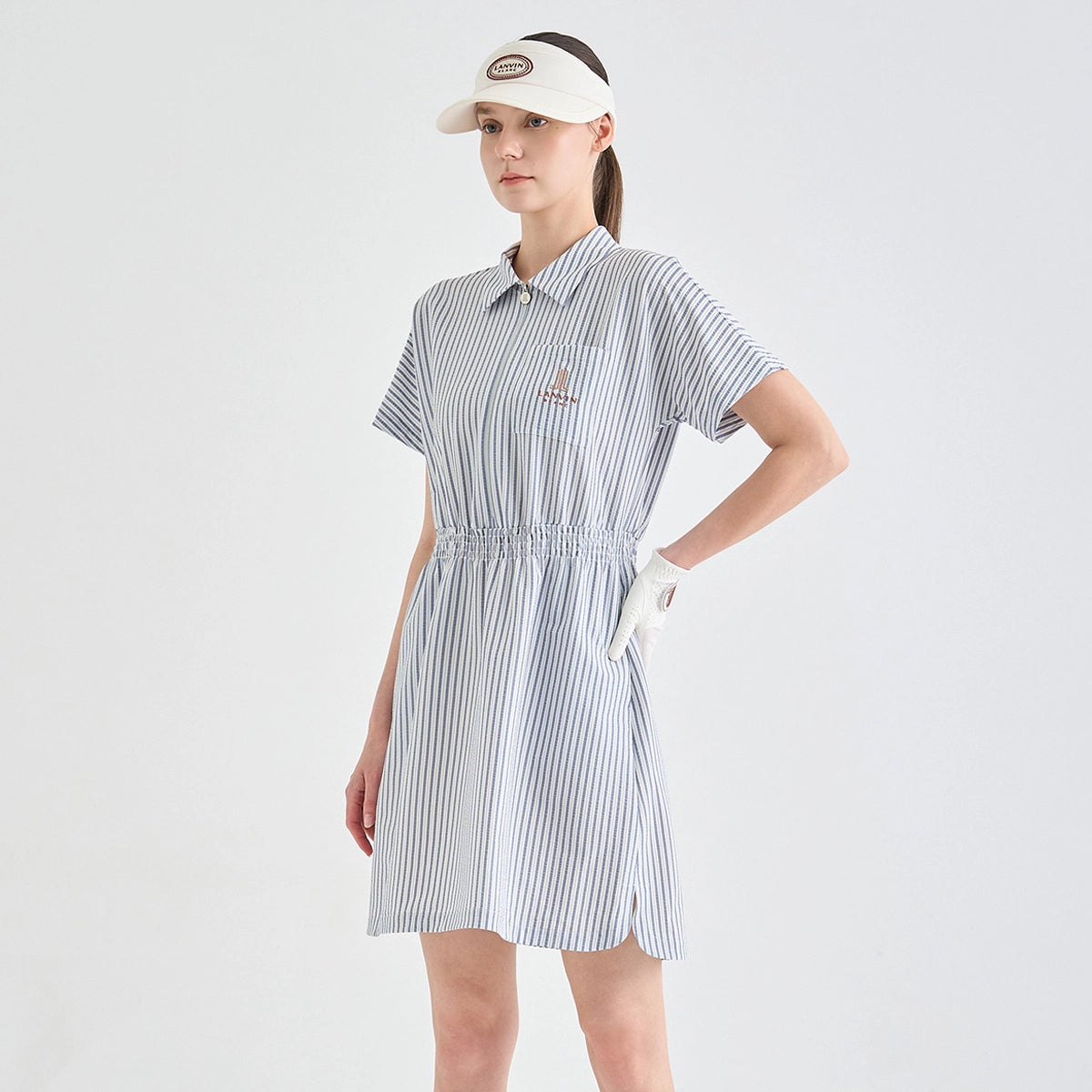 A woman modeling a short-sleeve collared golf dress with vertical blue and white stripes, featuring a drawstring waist and a small embroidered logo on the chest. She is also wearing a white visor and a white golf glove against a clean, light gray studio background.