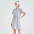 A woman modeling a short-sleeve collared golf dress with vertical blue and white stripes, featuring a drawstring waist and a small embroidered logo on the chest. She is also wearing a white visor and a white golf glove against a clean, light gray studio background.