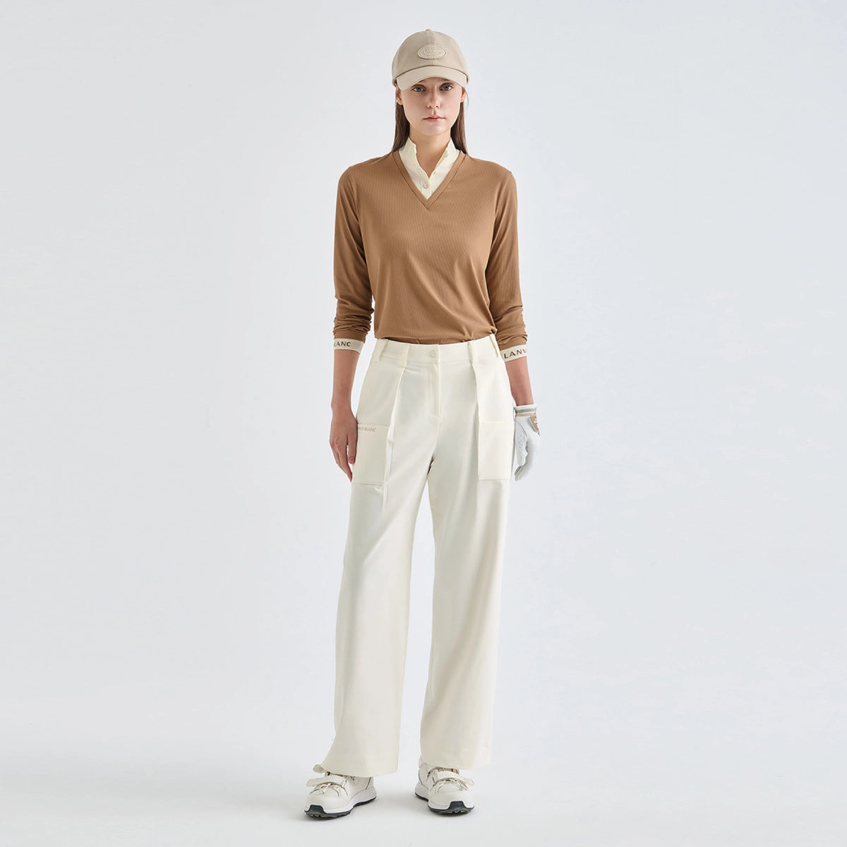 A model stands against a light gray studio background wearing a tan long-sleeve V-neck sweater with cream trim, off-white wide-leg trousers, a beige cap, white gloves, and white sneakers.