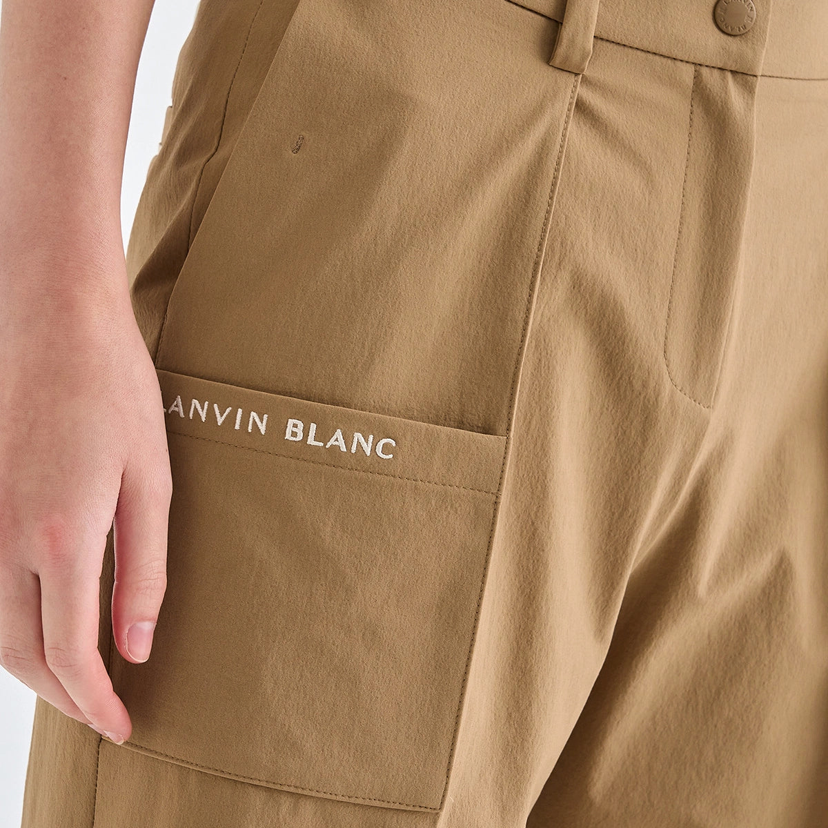 Close-up of a person wearing tan cargo-style pants, showing a front pocket with the text 'LANVIN BLANC' embroidered on the pocket, and part of the hand resting beside it.