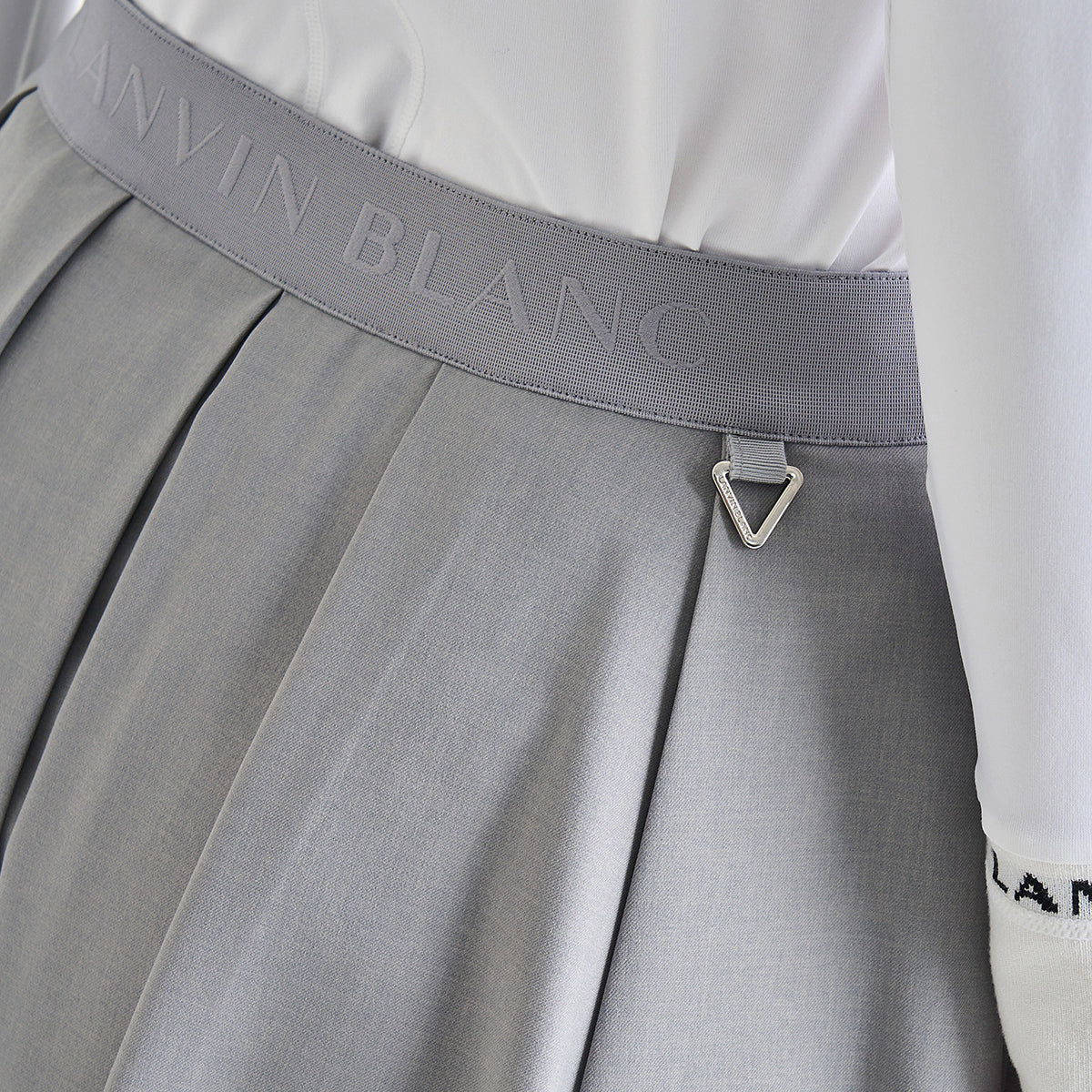 Close-up of a gray pleated skirt with a wide gray waistband embossed with the Calvin Klein logo, paired with a white top and a small silver triangular metal charm at the front.