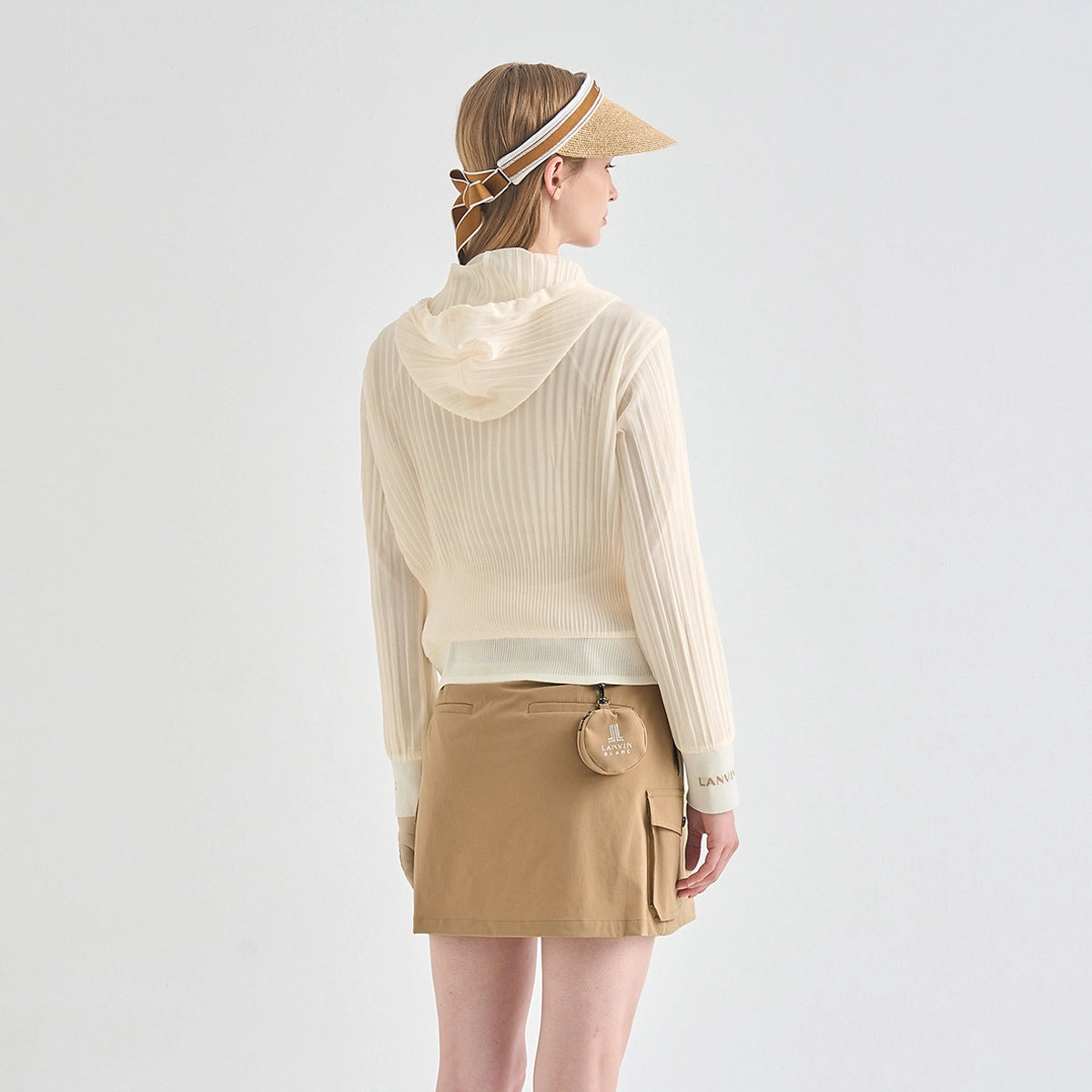 A woman shown from the back wearing a cream ribbed knit hooded long-sleeve top, a tan mini skirt with cargo pockets, a straw visor, and a patterned headscarf, against a plain light gray studio background.