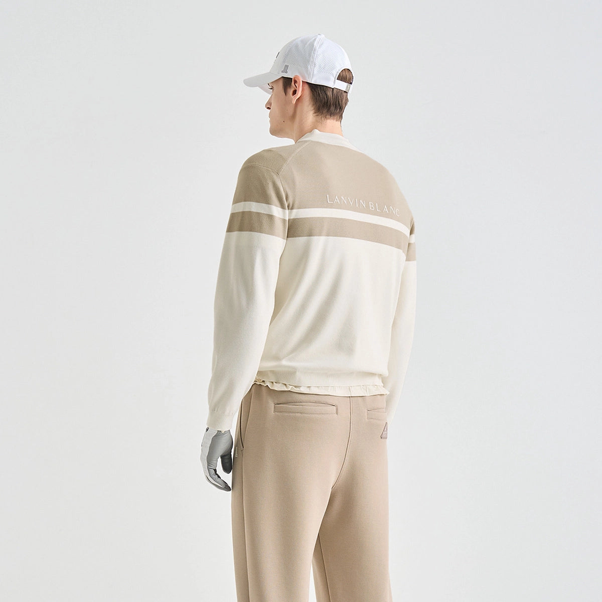 A man shown from the back wearing a beige and cream long-sleeve knit sweater with horizontal stripes, matching beige pants, a white baseball cap, and a gray glove, against a plain light gray studio background.