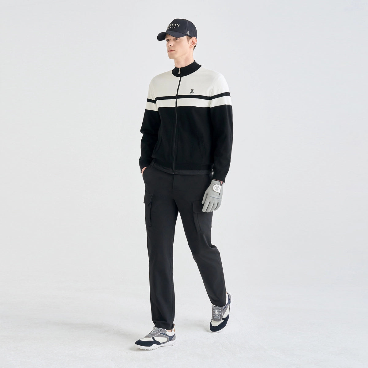 A man modeling a black and white color-blocked zip-up sweater, black pants, a black cap, and a gray golf glove against a light gray studio background.