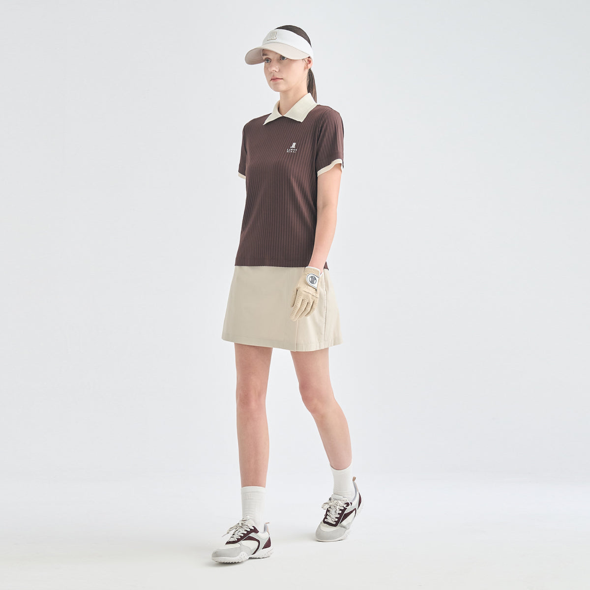 A woman modeling a dark brown short-sleeve polo shirt with a cream collar and sleeve trim, paired with a beige mini skirt, white visor, golf glove, white socks, and golf shoes against a light gray studio background.