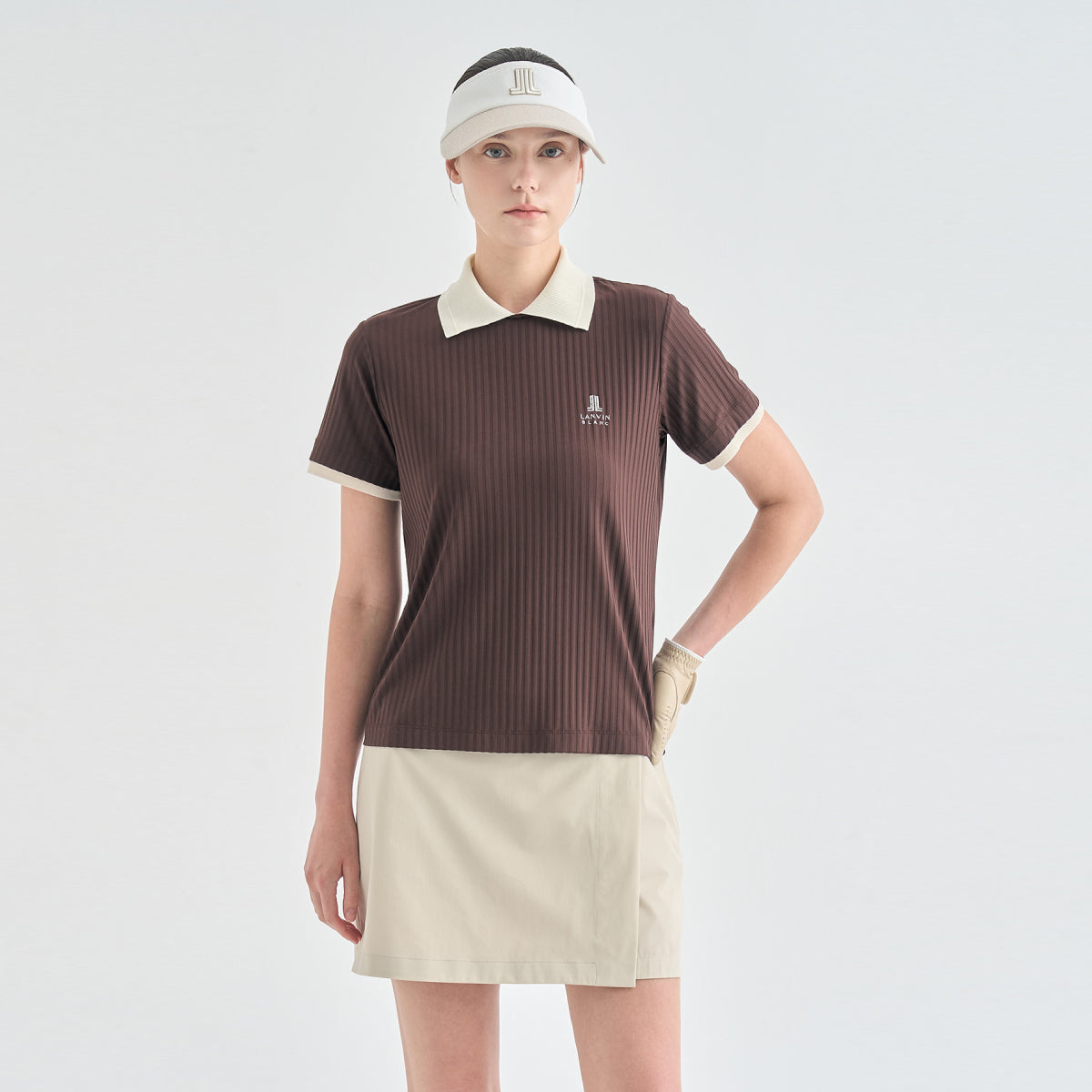 A woman modeling a ribbed short-sleeve polo shirt in dark brown with cream collar and sleeve trim, paired with a cream golf skirt, visor, and glove against a light gray studio background.