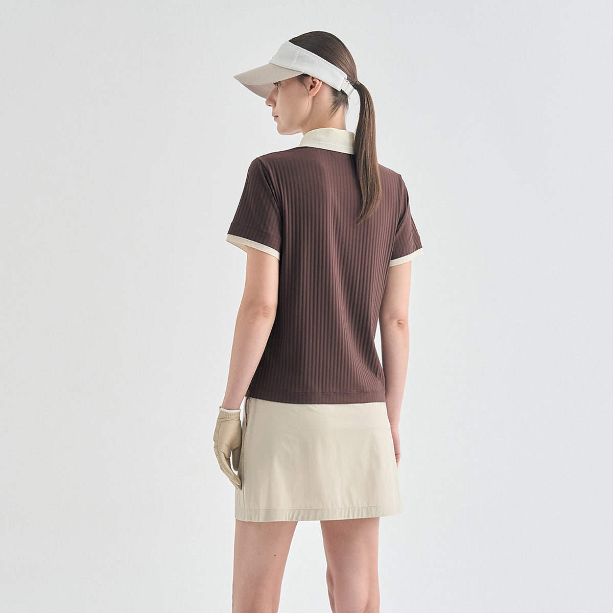 A woman shown from the back wearing a brown ribbed short-sleeve polo-style top with cream trim, a cream skirt, a white visor, and a white glove, against a plain light gray studio background.