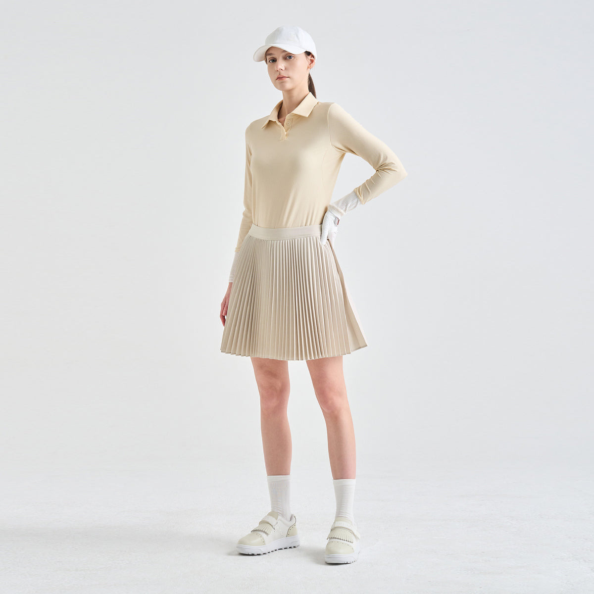 A woman modeling a beige long-sleeve collared knit top and matching pleated mini skirt, styled with a white cap, white gloves, white socks, and white sneakers against a light gray studio background.