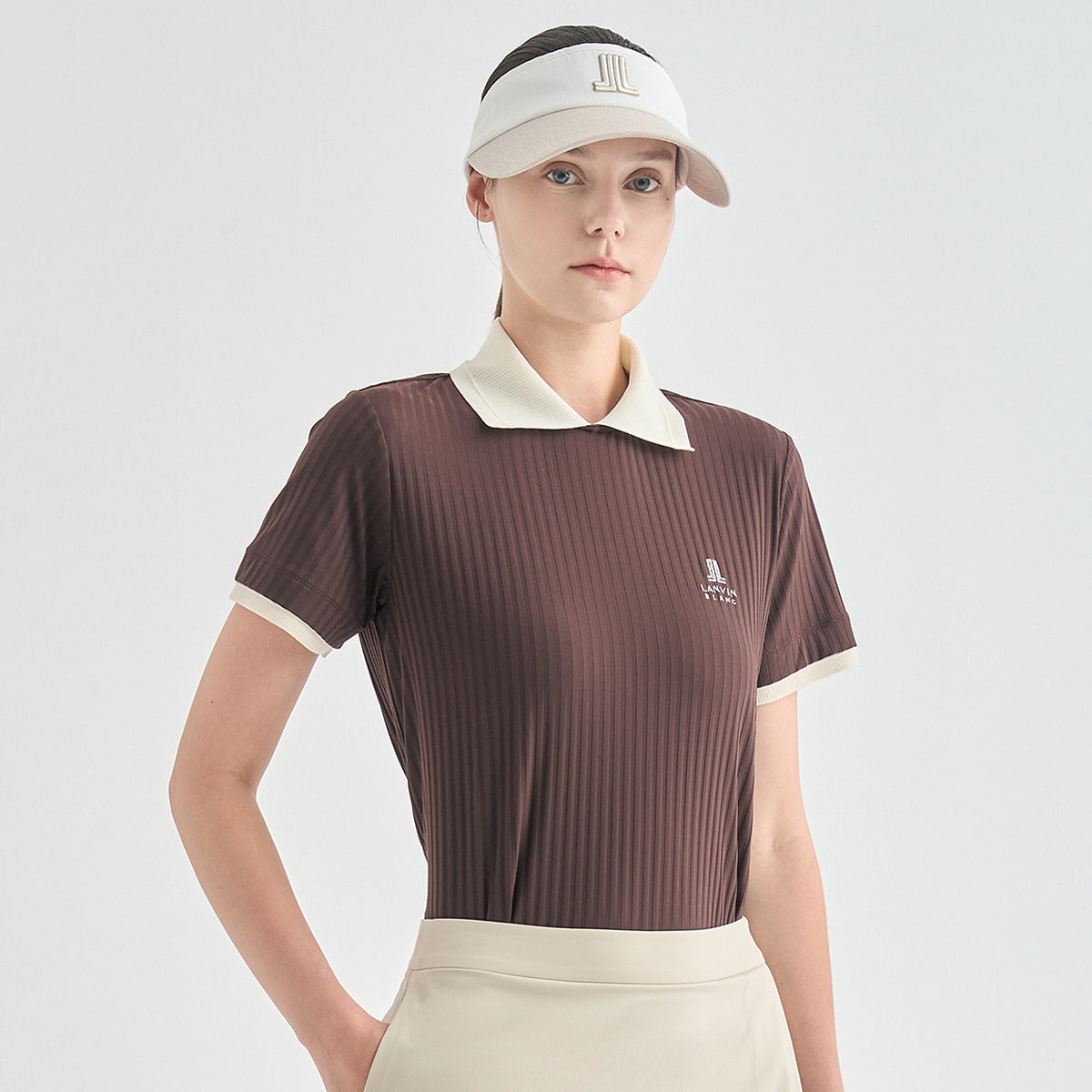 A model wearing a short-sleeve ribbed polo shirt in dark brown with cream accents on the collar and sleeve cuffs, paired with a matching cream visor and skirt, against a light gray studio background.