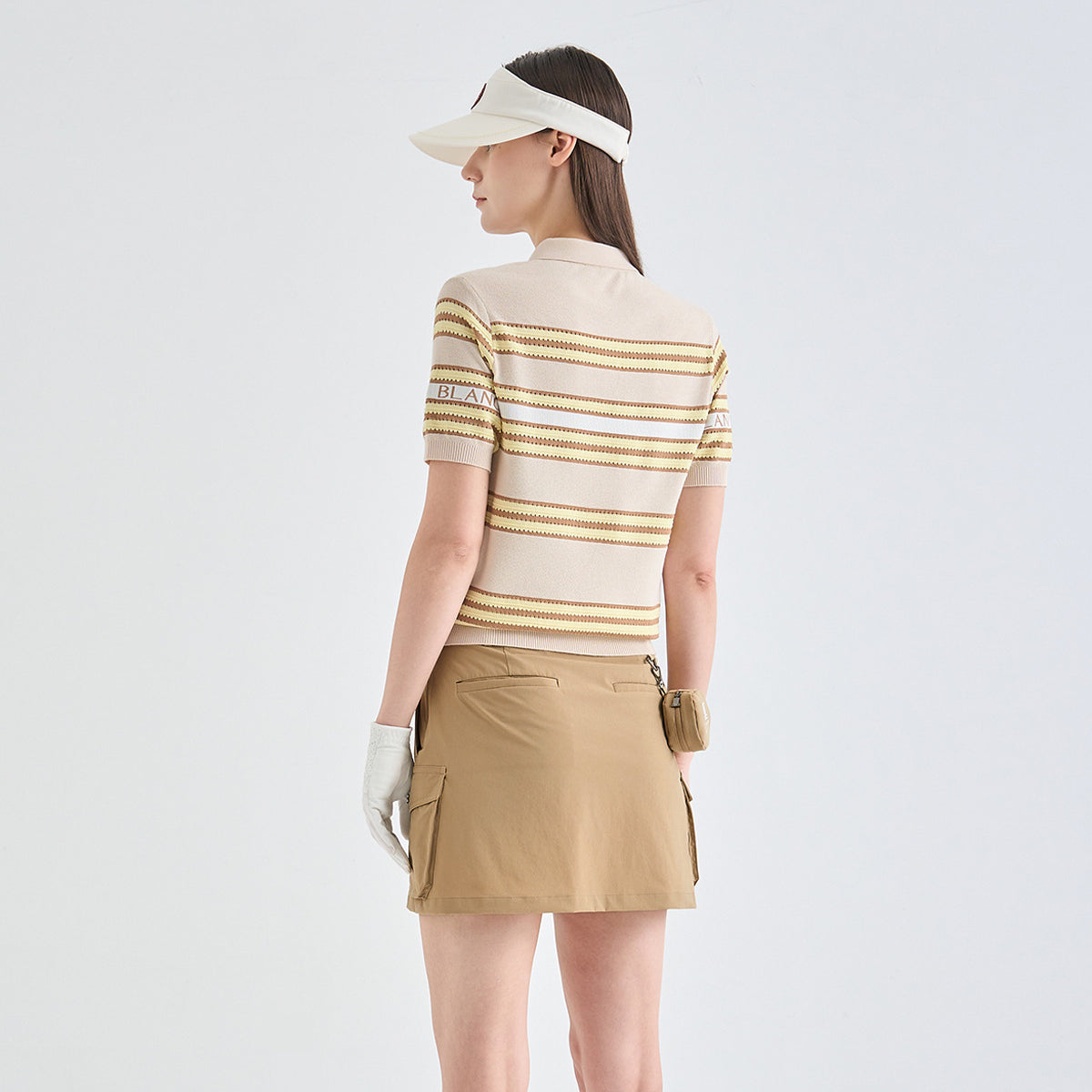 A woman is shown from the back wearing a beige and gold striped short-sleeve polo shirt, a tan cargo mini skirt, a white visor, and a white glove. The brand name 'LANVIN BLANC' is visible on the sleeve.
