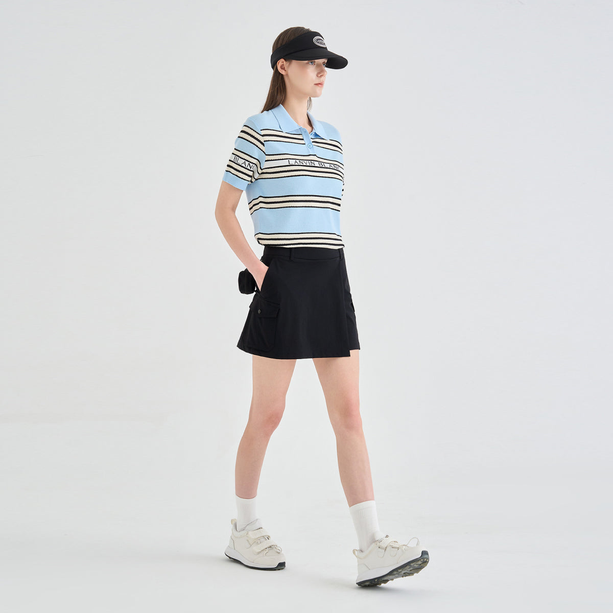 A model wearing a light blue and black striped short-sleeve polo shirt, a black mini skirt, white socks, and white sneakers, standing against a light gray studio background.
