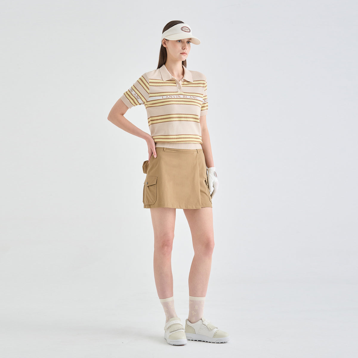 A model stands against a light gray studio background wearing a beige and white striped short-sleeve polo shirt, a tan cargo mini skirt, a beige visor, white golf gloves, white socks, and white sneakers.