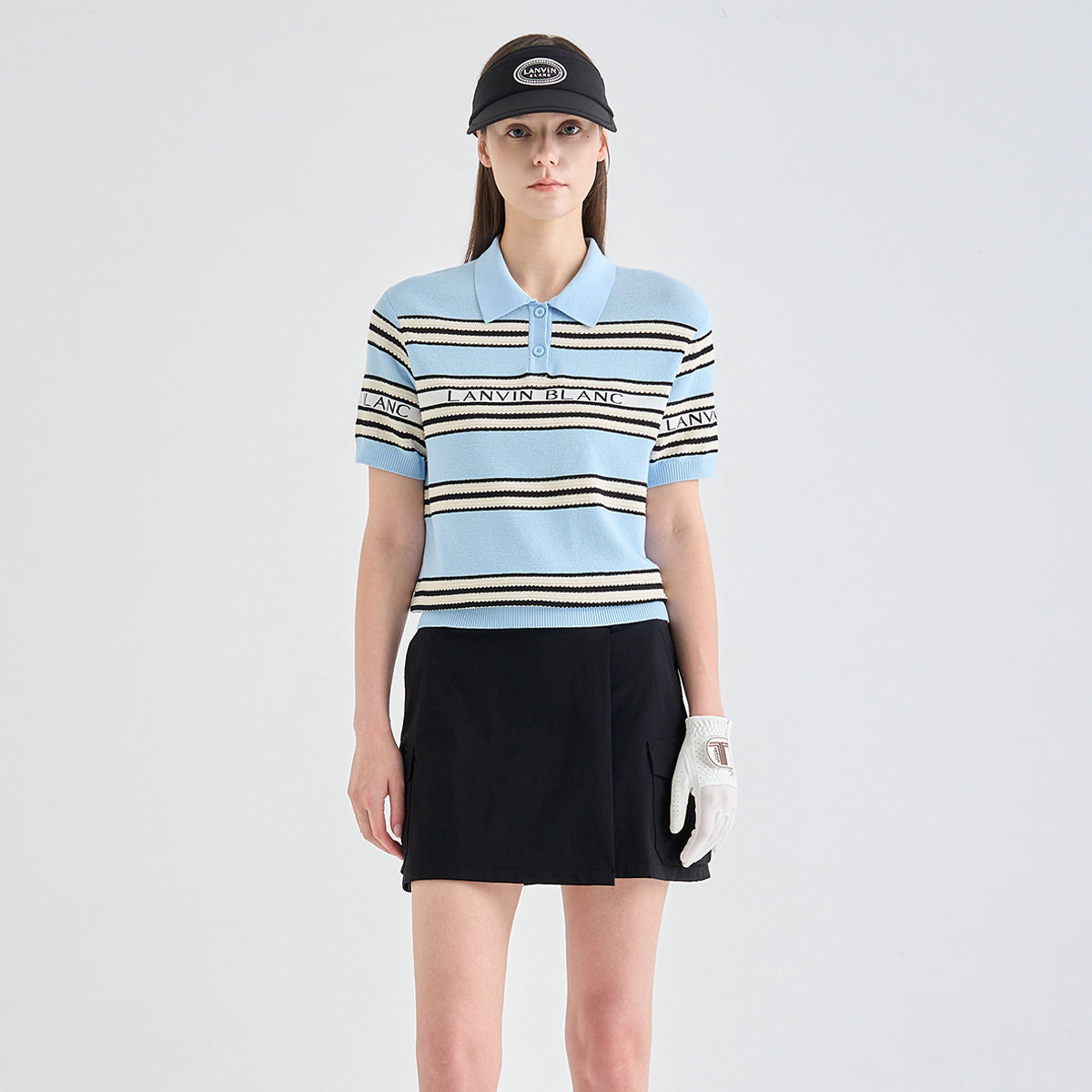 A model stands against a light gray studio background wearing a light blue short-sleeve polo shirt with black and white horizontal stripes, a black mini skirt, a black visor, and a white golf glove.