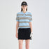 A model stands against a light gray studio background wearing a light blue short-sleeve polo shirt with black and white horizontal stripes, a black mini skirt, a black visor, and a white golf glove.