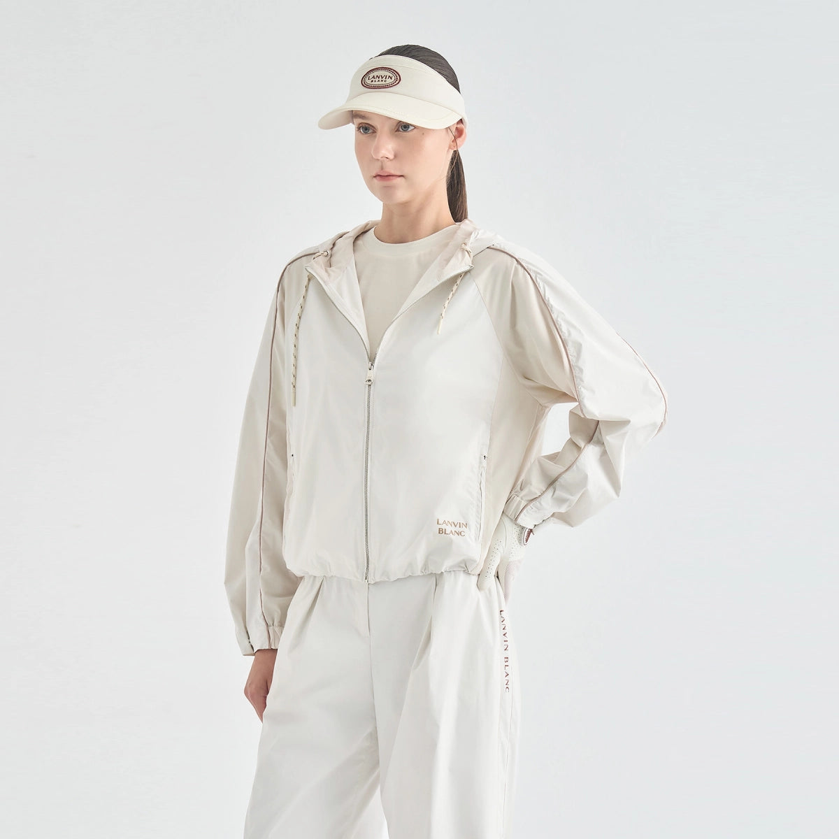 A model wearing a light beige hooded windbreaker jacket, matching beige pants, a beige visor with a circular logo patch, and a beige crewneck top, posed against a solid light gray studio background.