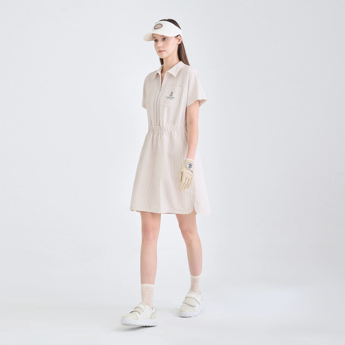 A woman stands in a studio wearing a short-sleeve collared golf dress with thin vertical stripes, a white visor, a white golf glove, white socks, and white sneakers. The background is a clean, light gray studio backdrop.