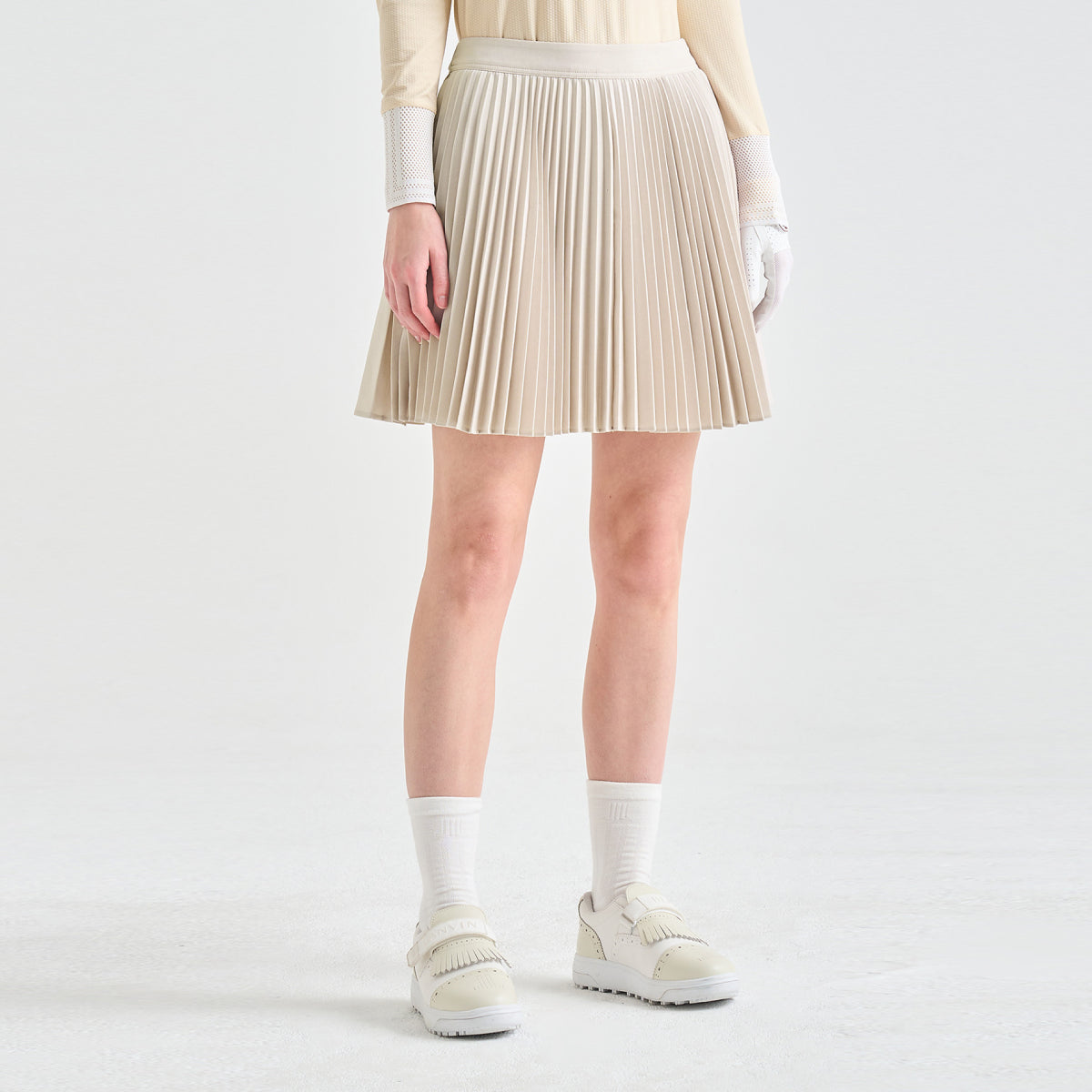 A model wearing a pleated beige mini skirt, cream knit long-sleeve top, white gloves, white socks, and cream platform sneakers, shown from the waist down against a light gray studio background.