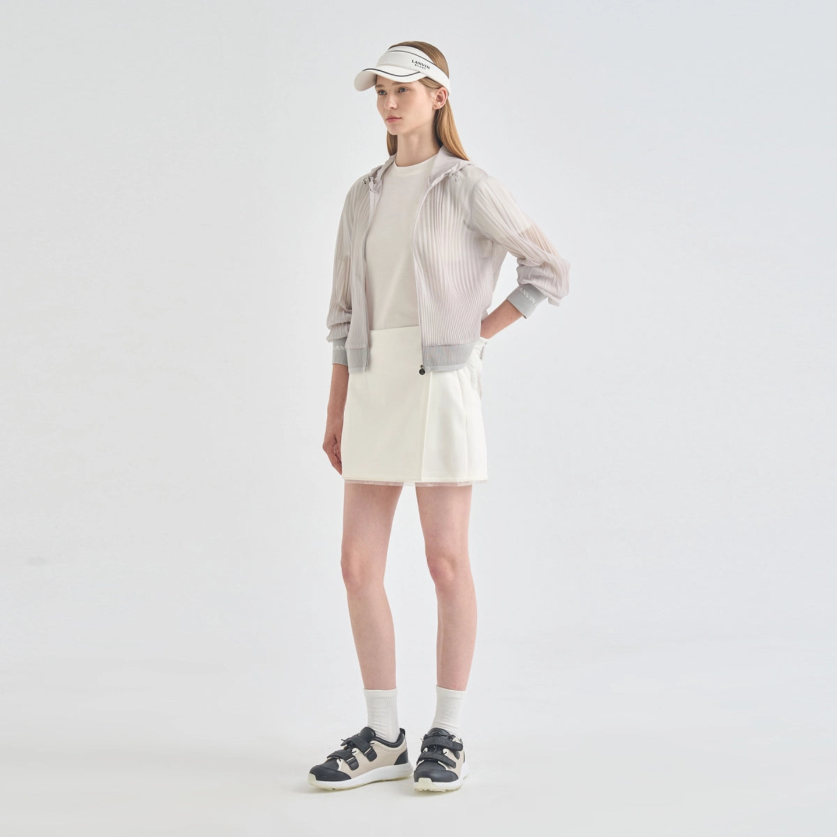 A model stands in a studio wearing a light beige and white athletic outfit, featuring a sheer long-sleeve zip-up hoodie over a white top, a white mini skirt, white socks, and black-and-white sneakers.