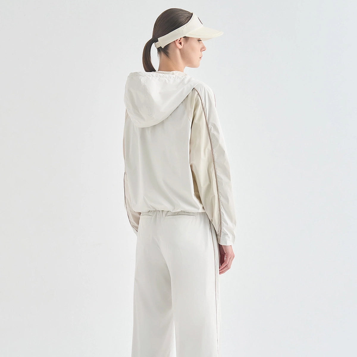A woman shown from the back wearing a light beige hooded windbreaker jacket with contrasting sleeves, matching light beige pants, and a beige visor against a plain white studio background.