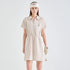 A woman stands against a light gray studio background wearing a short-sleeve collared golf dress with a front zipper, a matching visor, and a glove. The dress has thin vertical stripes in off-white and beige and a small embroidered logo on the left chest.