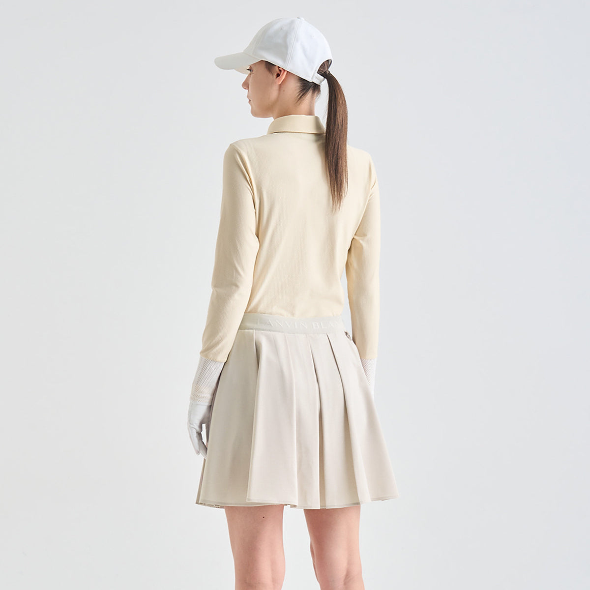 A woman shown from the back wearing a light beige long-sleeve mock-neck top, a matching pleated mini skirt, white gloves, and a white baseball cap against a plain light gray studio background.