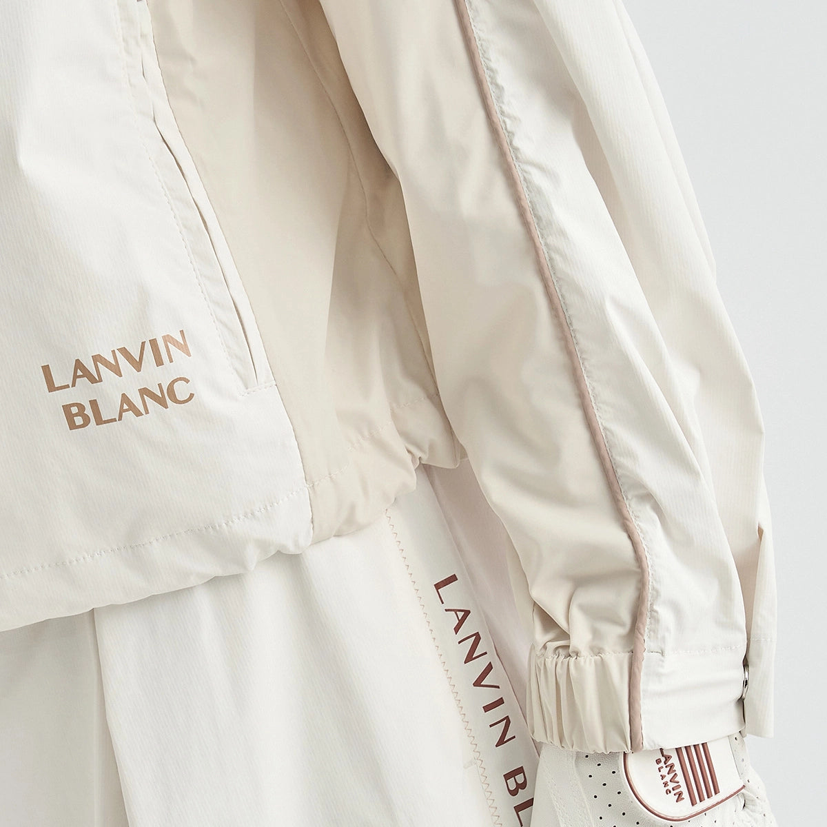 Close-up of a white Lanvin Blanc lightweight jacket with gold-toned 'LANVIN BLANC' text on the chest and sleeve, plus a white sneaker partially visible underneath.