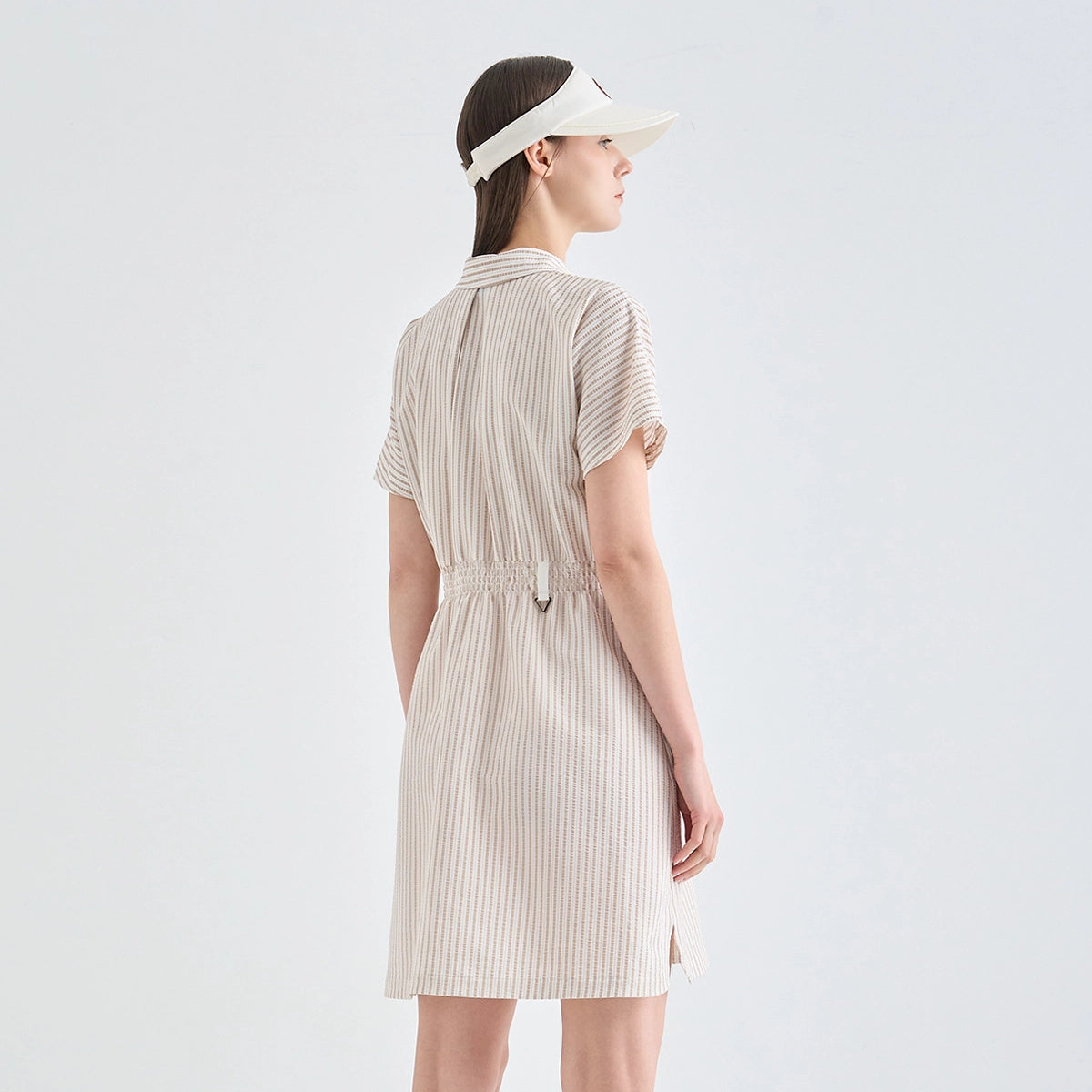 A woman shown from the back wearing a short-sleeve beige and white vertically striped shirt dress with a tie waist, paired with a matching visor, against a light gray studio background.