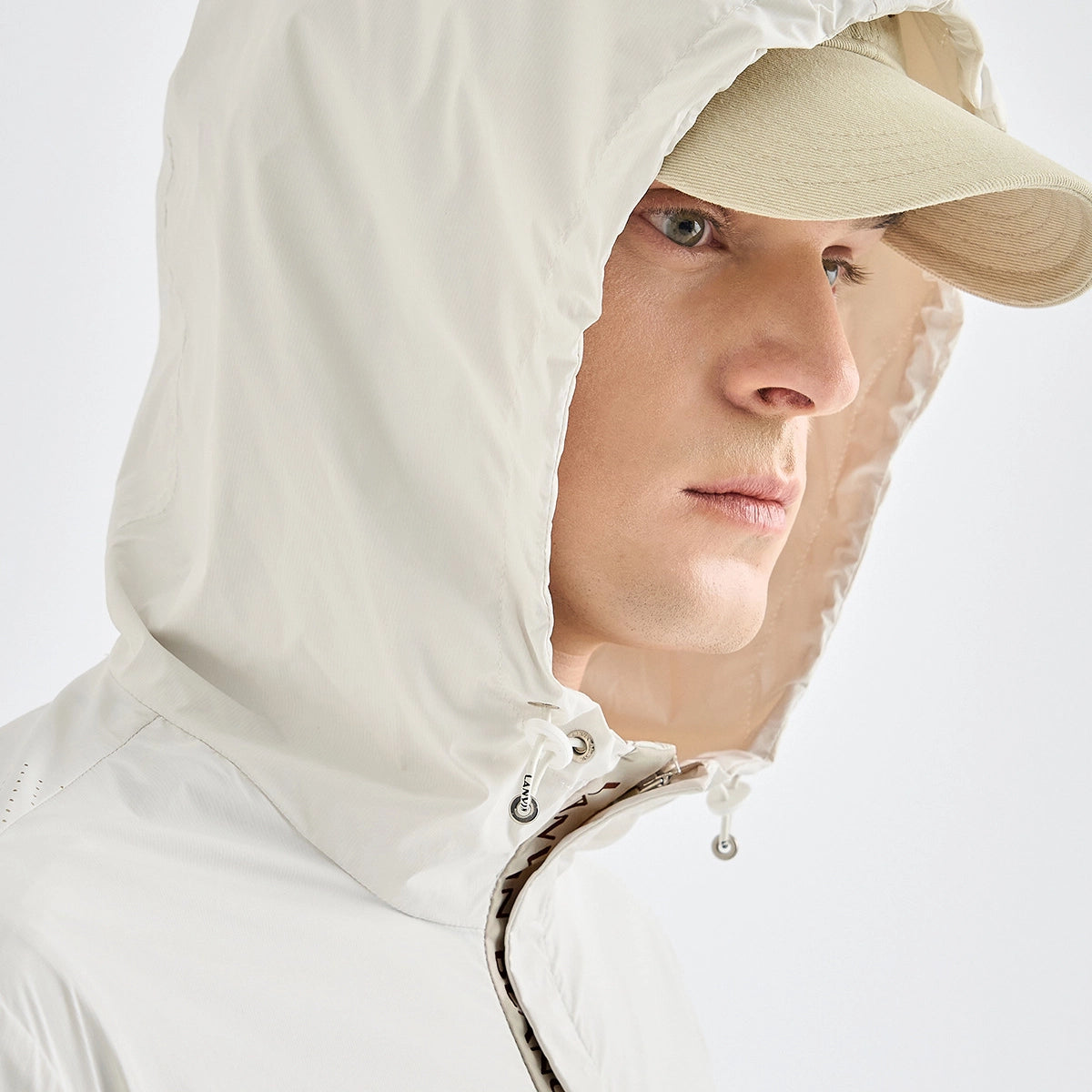 Close-up of a person wearing a light off-white hooded jacket with drawstrings and a beige baseball cap against a clean white studio background.