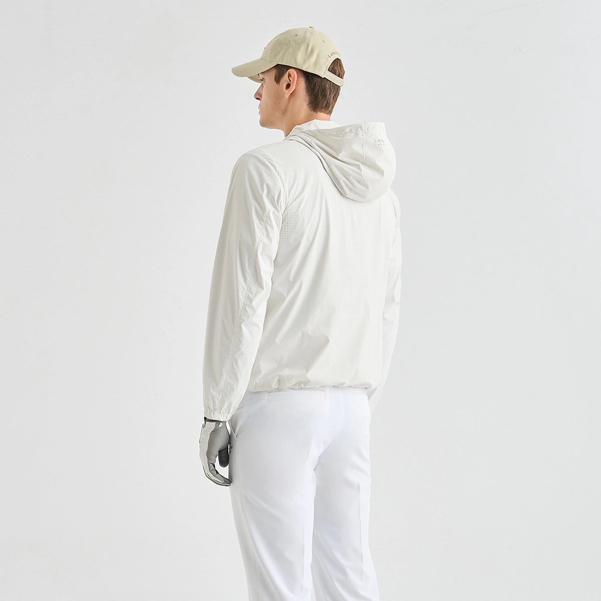 A model shown from the back wearing a light off-white hooded windbreaker jacket, white pants, a beige baseball cap, and a silver glove, set against a plain light gray studio background.
