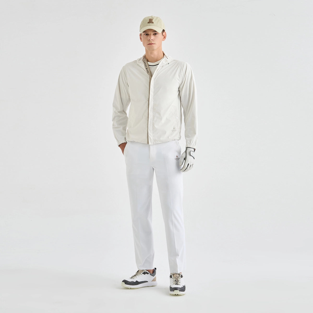 A male model stands against a plain white studio background wearing a light beige zip-up jacket, white golf pants, a beige cap, a white golf glove, and white and black golf shoes.