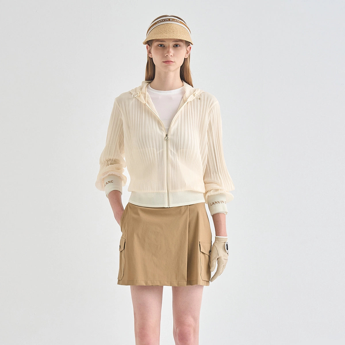 A woman stands against a light gray studio background wearing a cream ribbed zip-front hooded jacket over a white top, a tan skirt, a beige visor, and a light glove. The jacket has subtle text on the cuffs and a small gold zipper pull.