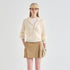 A woman stands against a light gray studio background wearing a cream ribbed zip-front hooded jacket over a white top, a tan skirt, a beige visor, and a light glove. The jacket has subtle text on the cuffs and a small gold zipper pull.