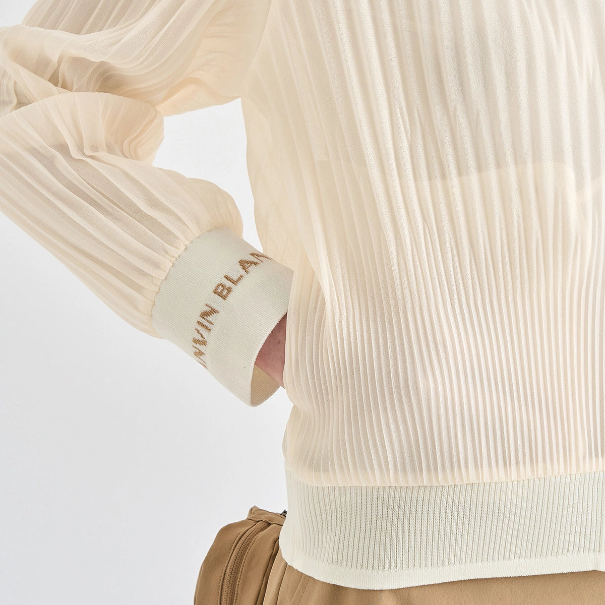 Close-up of a person wearing a cream-colored pleated long-sleeve top with a ribbed cuff featuring the text 'ALVIN BLA.