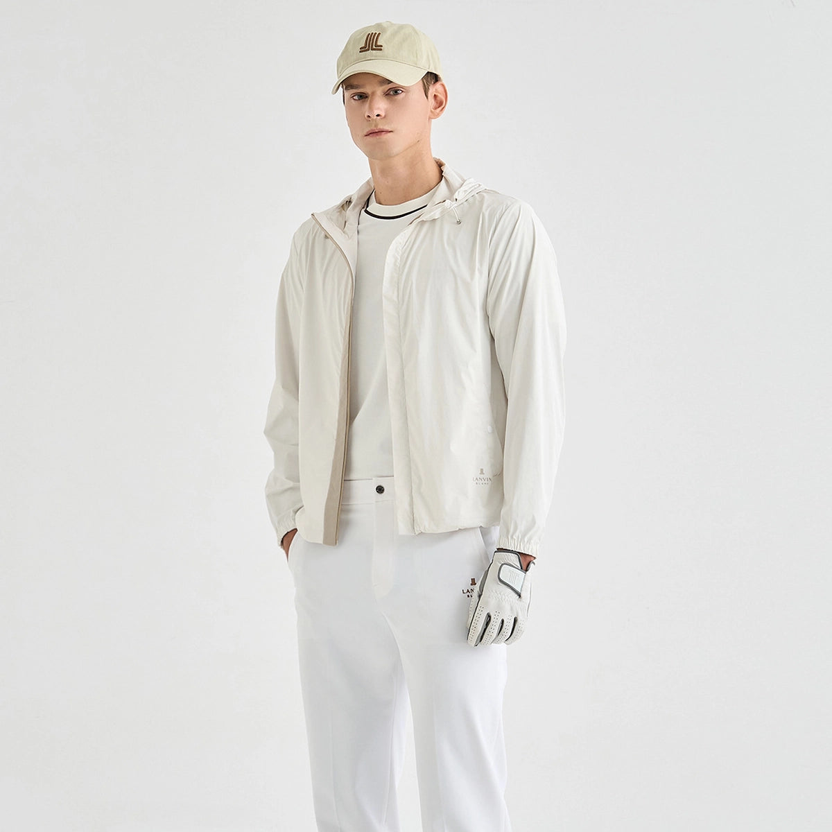 A man modeling a light beige outdoor jacket, white pants, a beige baseball cap, and a white glove against a plain white studio background.