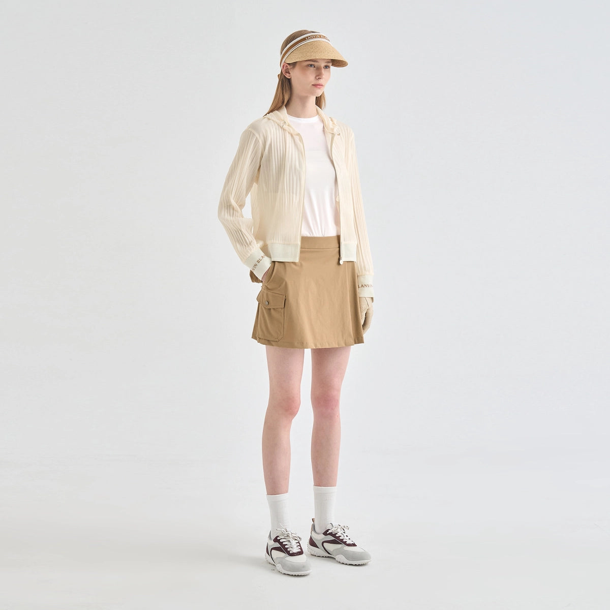 A model stands against a light gray studio background wearing a beige visor, a cream pleated lightweight jacket over a white top, a tan mini skirt with side pockets, white socks, and gray sneakers.