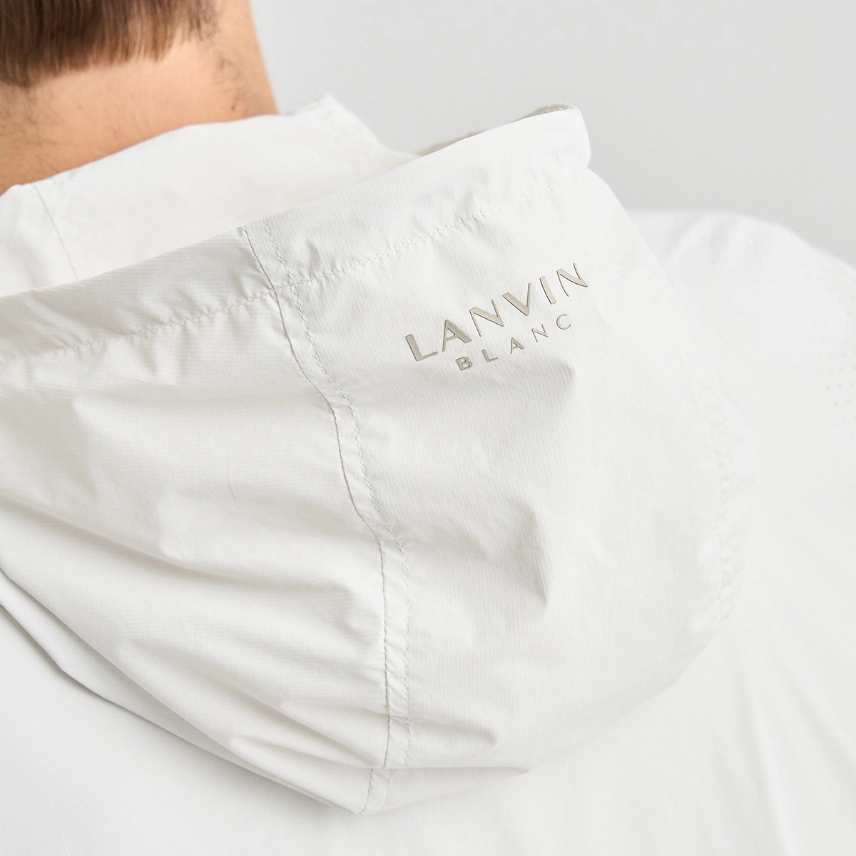 Close-up of a white hooded jacket with the brand name 'LANVIN BLANC' printed in a subtle metallic tone on the side of the hood, worn by a model.