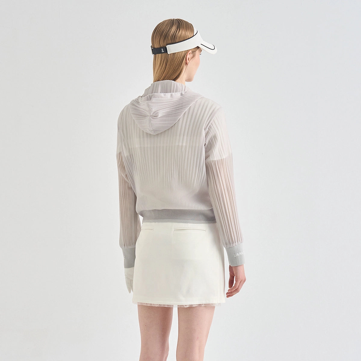 A woman shown from the back wearing a light beige ribbed long-sleeve hooded top with sheer mesh sleeves, a white visor, and a white mini skirt against a plain light gray studio background.