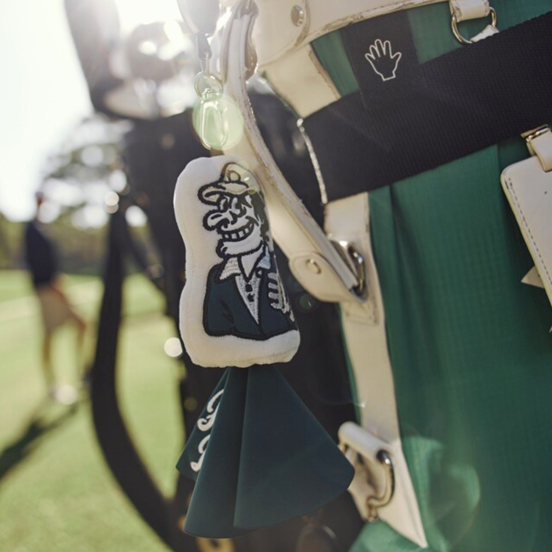 A close-up lifestyle shot of a golf bag with a plush golf ball tag featuring a skeleton character in a suit, hanging from a black strap on a green golf course.