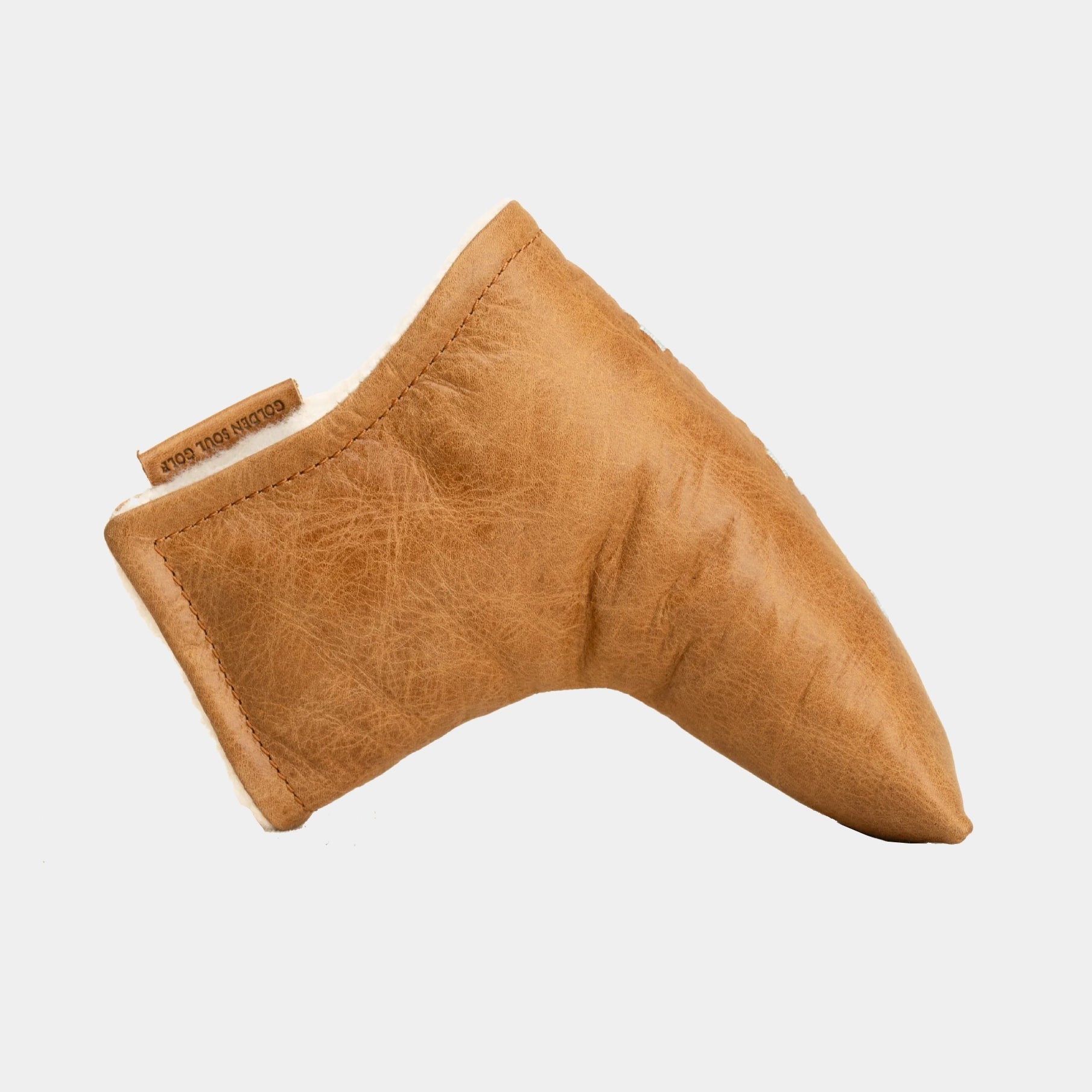 A tan leather blade putter cover with a small brown leather brand tag reading 'cook and mange' on a light gray background.