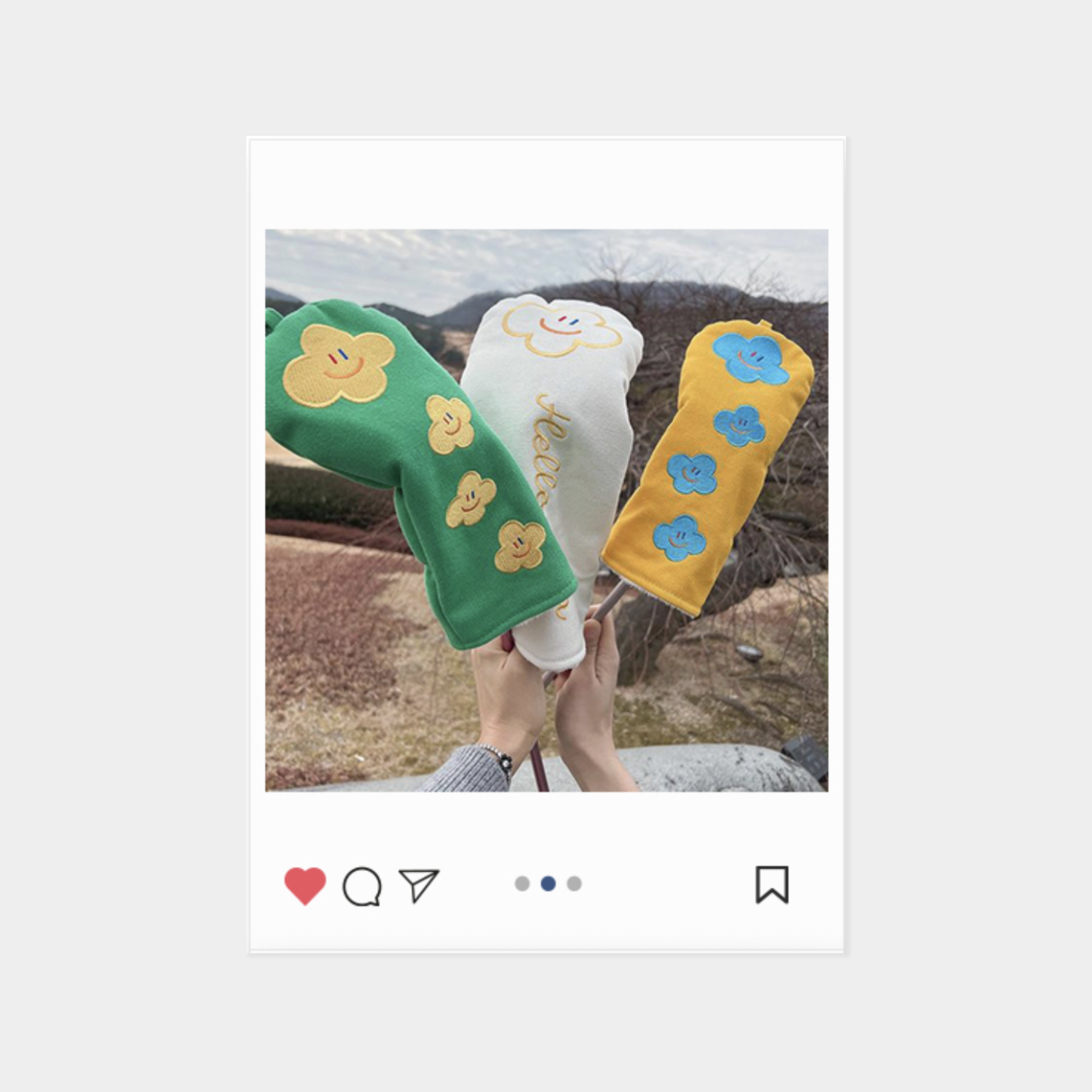 Three colorful knit sleeve-like headcovers with embroidered flower or cloud motifs are held up outdoors. One is green with yellow flowers, one is white with a yellow cloud and smiley face, and one is yellow with blue flowers.