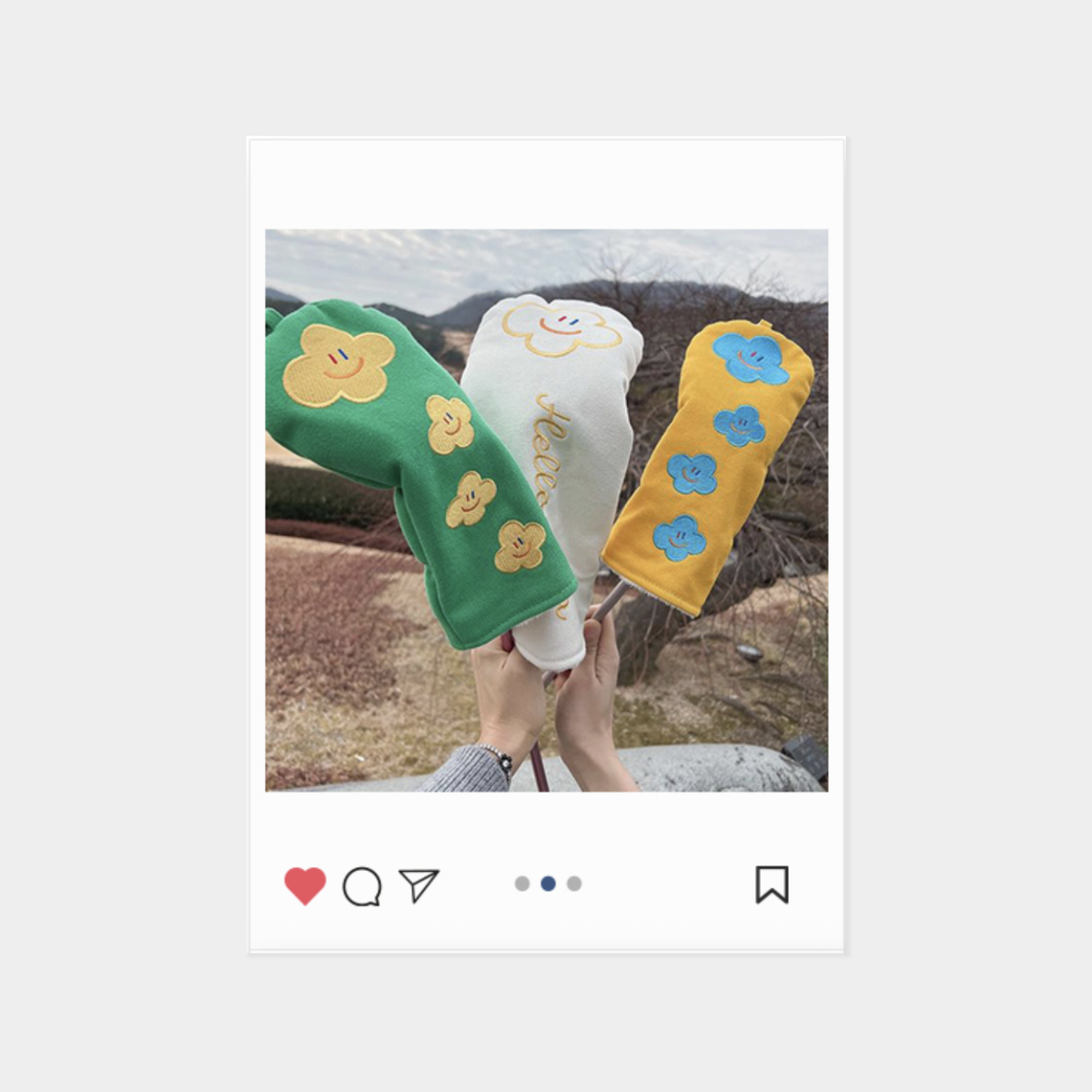 Three colorful knit sleeve-like headcovers with embroidered flower or cloud motifs are held up outdoors. One is green with yellow flowers, one is white with a yellow cloud and smiley face, and one is yellow with blue flowers.