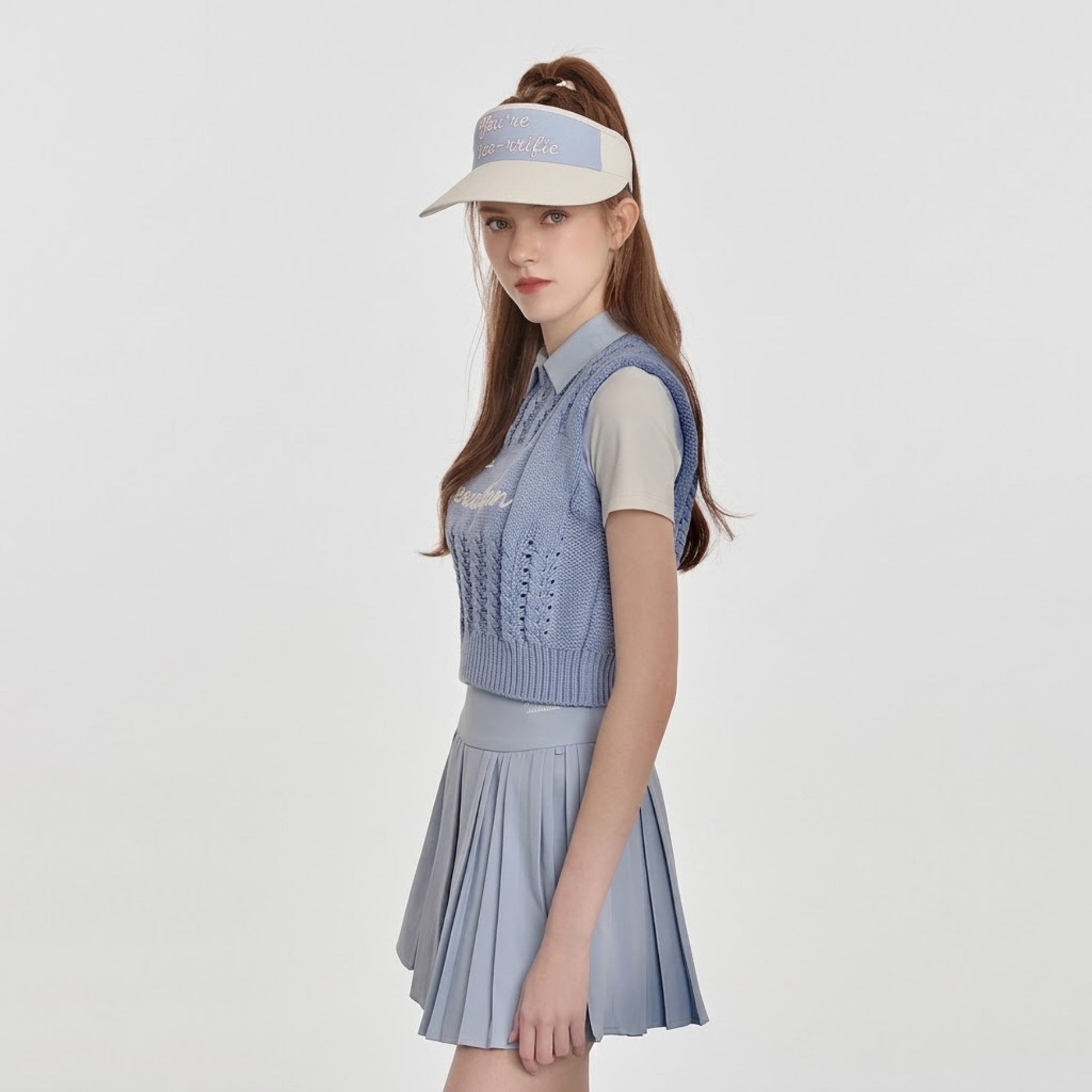 A model wearing a light blue knit polo vest over a white short-sleeve collared shirt, paired with a matching light blue pleated mini skirt and a beige visor, set against a plain light gray background.