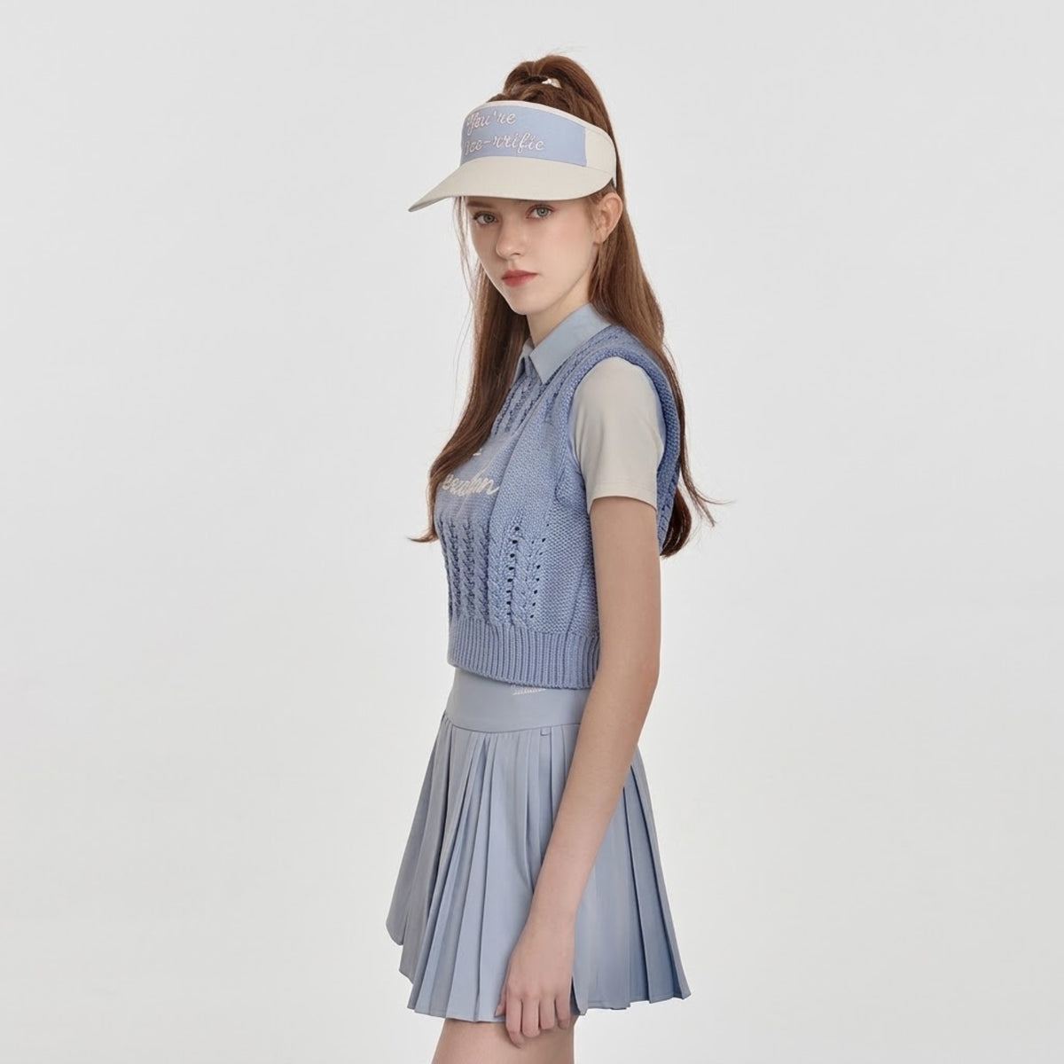 A model wearing a light blue knit polo vest over a white short-sleeve collared shirt, paired with a matching light blue pleated mini skirt and a beige visor, set against a plain light gray background.